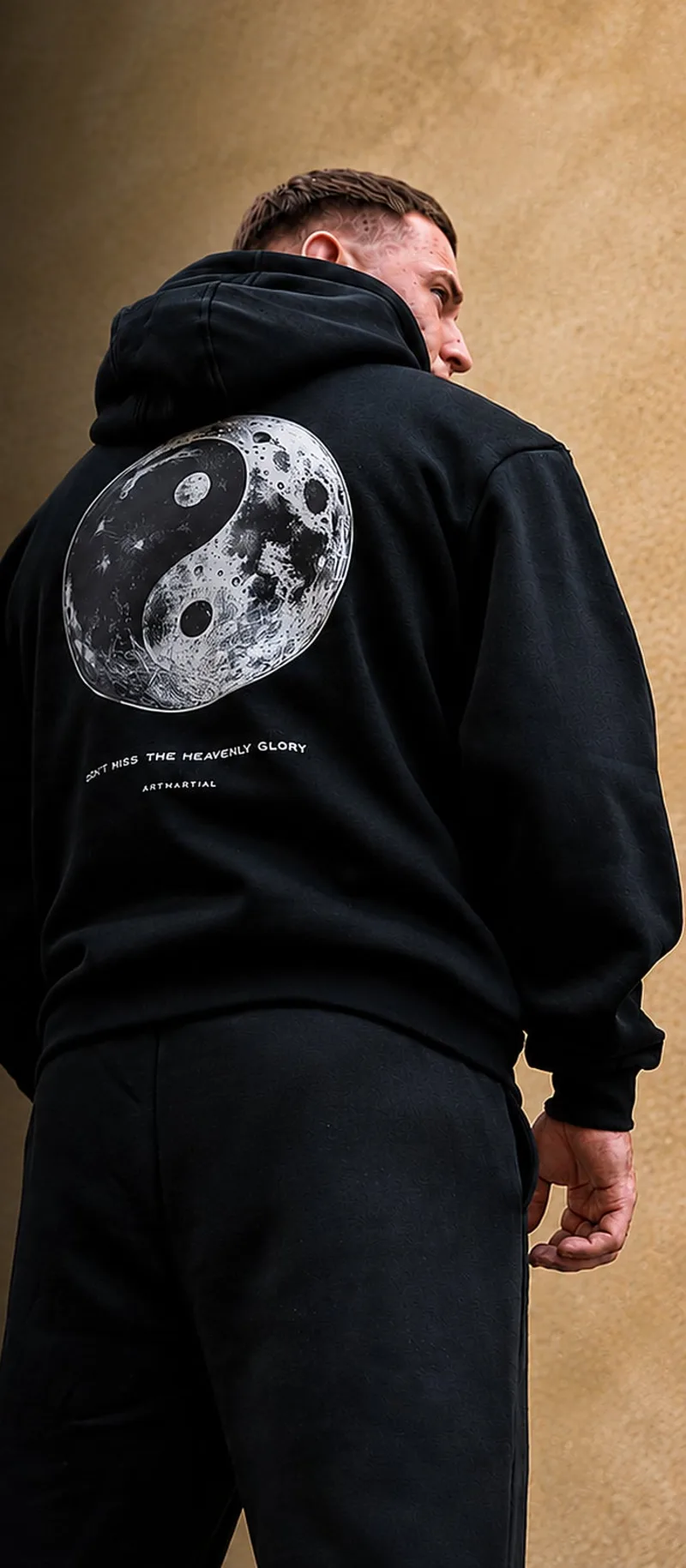 A man in a black ArtMartial hoodie with a moon yin-yang print, seen from behind against a sandstone wall