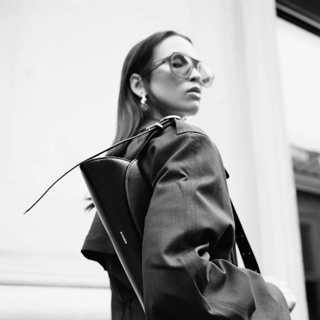 A woman in tinted glasses and a dark coat with a black leather bag over her shoulder, in black and white