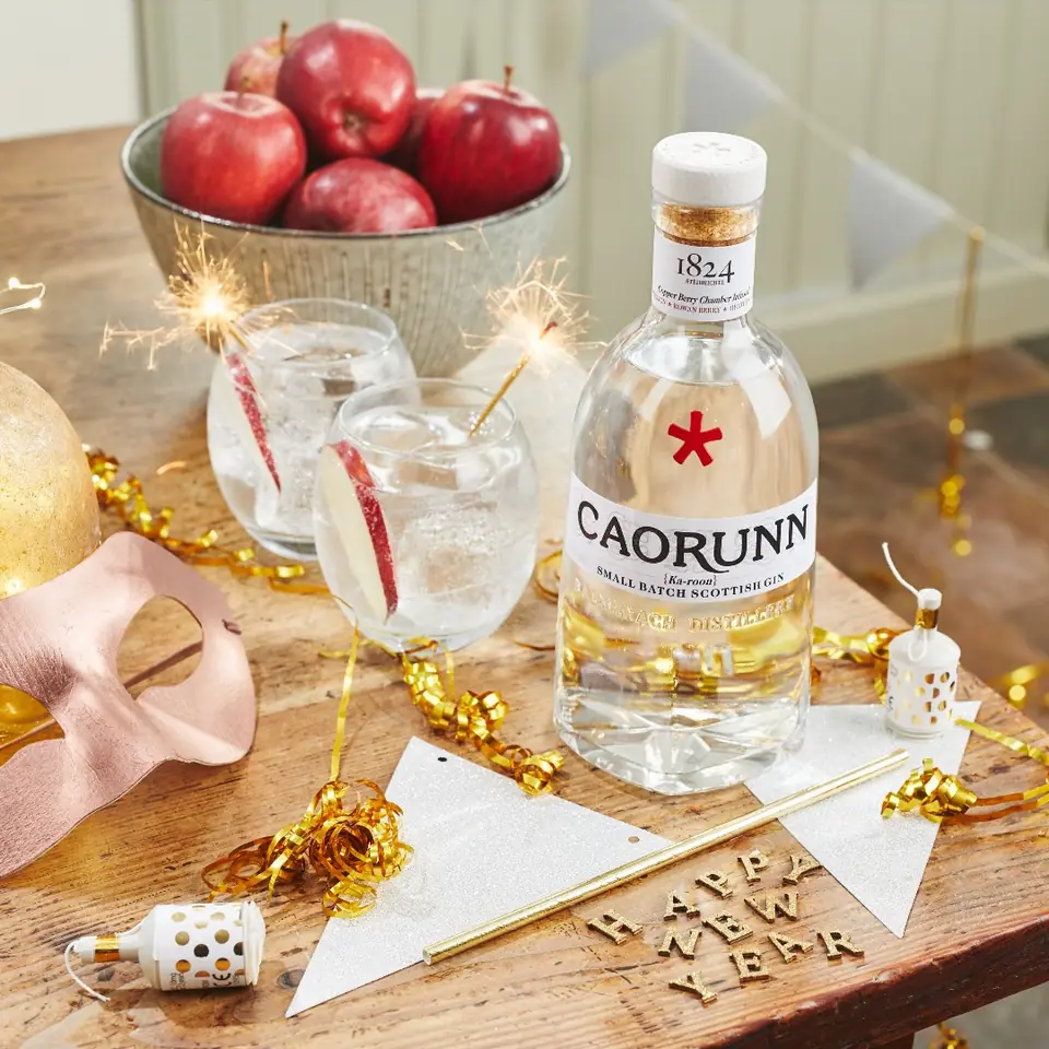 A bottle of Caorunn gin on a table with red apples and a gin and tonic