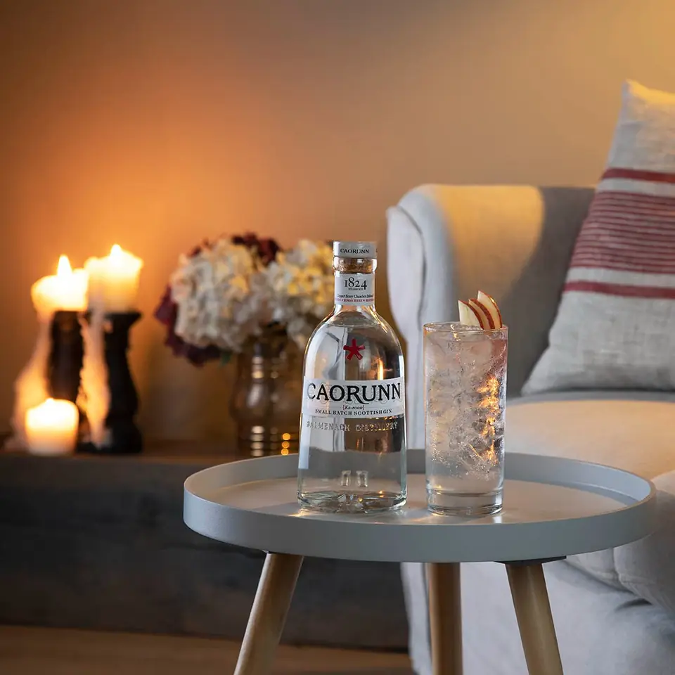 A bottle of Caorunn gin and a glass on a side table by candlelight