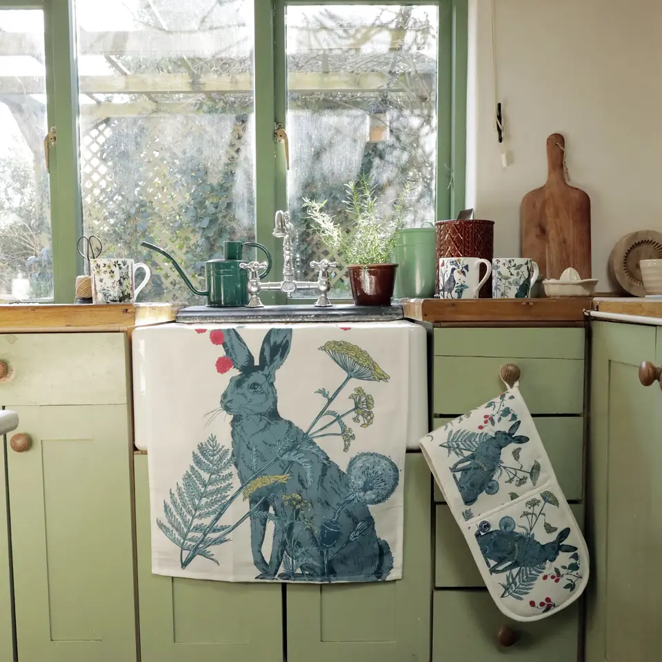 A hare-print tea towel and oven glove hanging in a green country kitchen
