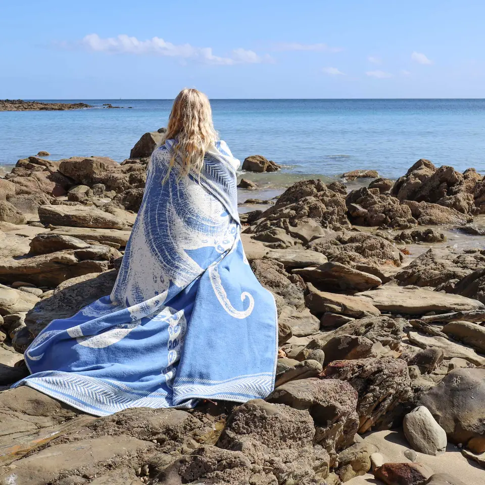 A woman in a blue octopus-print Cream Cornwall throw, sitting on rocks above a Cornish beach