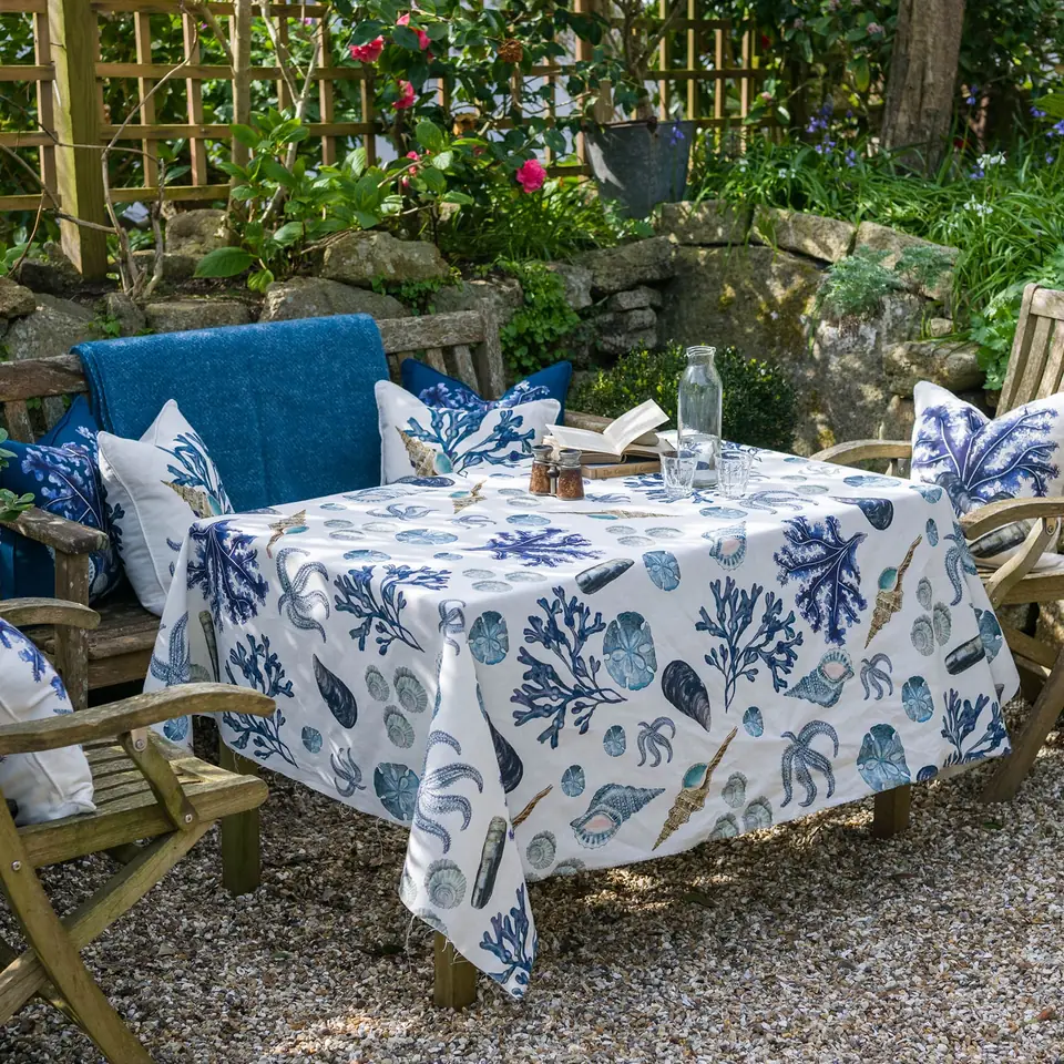 A garden table dressed in a blue and white rockpool-print linen cloth, with cushions on the bench beside it