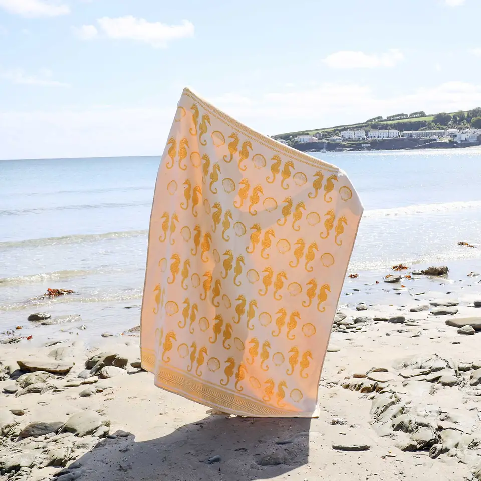 A golden yellow seahorse-print towel held up on a sandy beach, the sea behind it