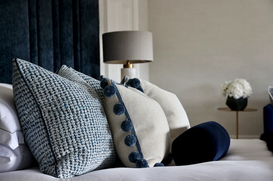 Navy pom-pom and woven cushions on a bed beside a table lamp