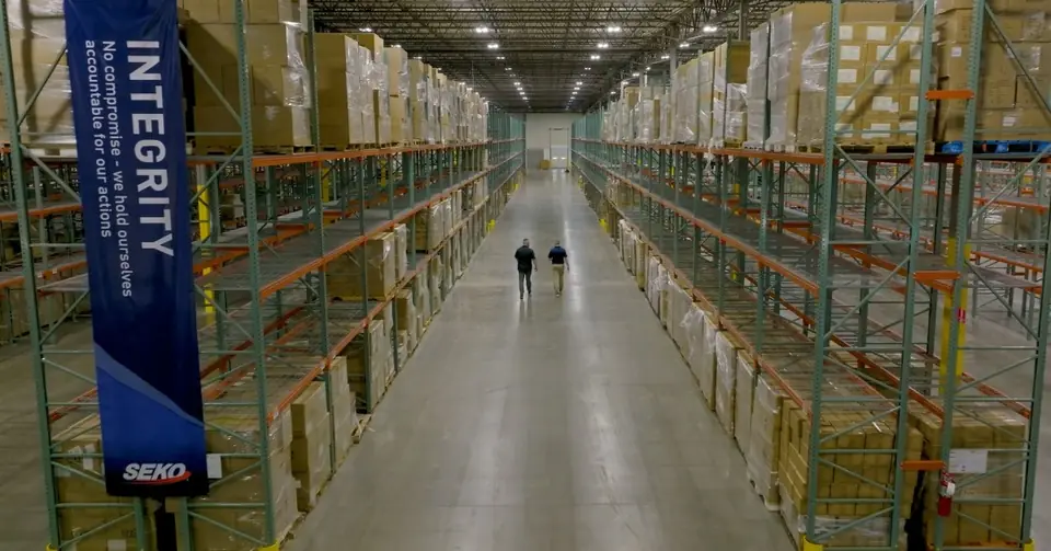 A long warehouse aisle of racked stock with two people walking through it