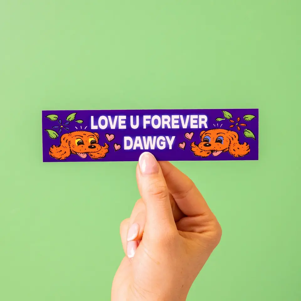 A hand holding a purple Love U Forever Dawgy bumper sticker against green
