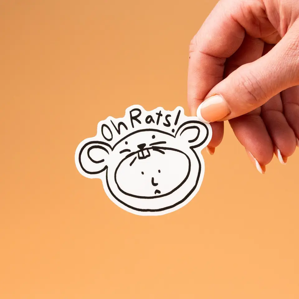 A hand holding an Oh Rats die-cut sticker against orange