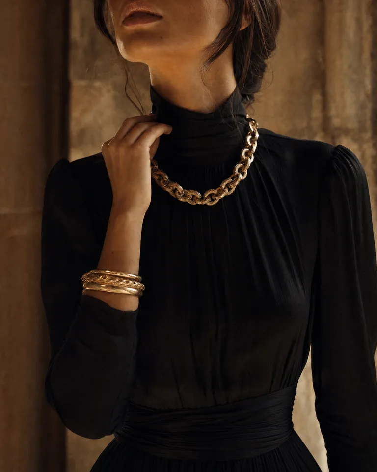 A woman in a black high-neck dress wearing a chunky gold chain and stacked gold bangles