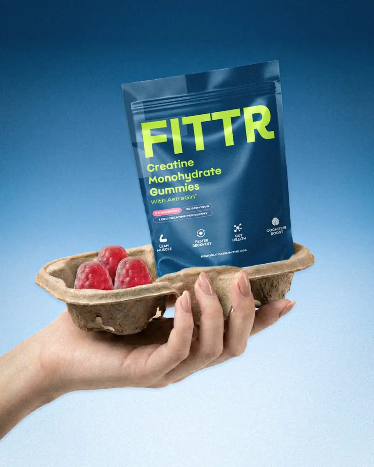 A hand holding a pouch of Fittr creatine gummies on a tray of berries against a blue backdrop