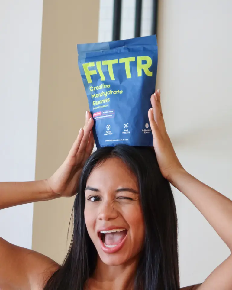 A smiling woman winking with a pouch of Fittr creatine gummies balanced on her head