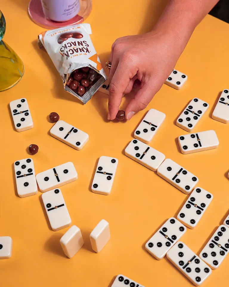 A hand reaching for Knack Snacks protein balls among dominoes on an orange table