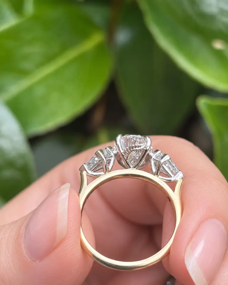 A hand holding a three-stone diamond engagement ring against green leaves