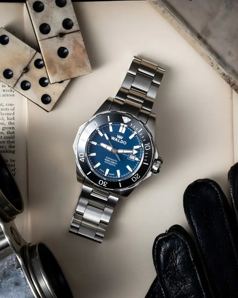 A Waldo watch with a blue dial laid among dominoes and leather gloves
