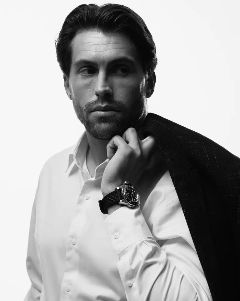 A man in a white shirt with a jacket over his shoulder wearing a Waldo watch, in black and white