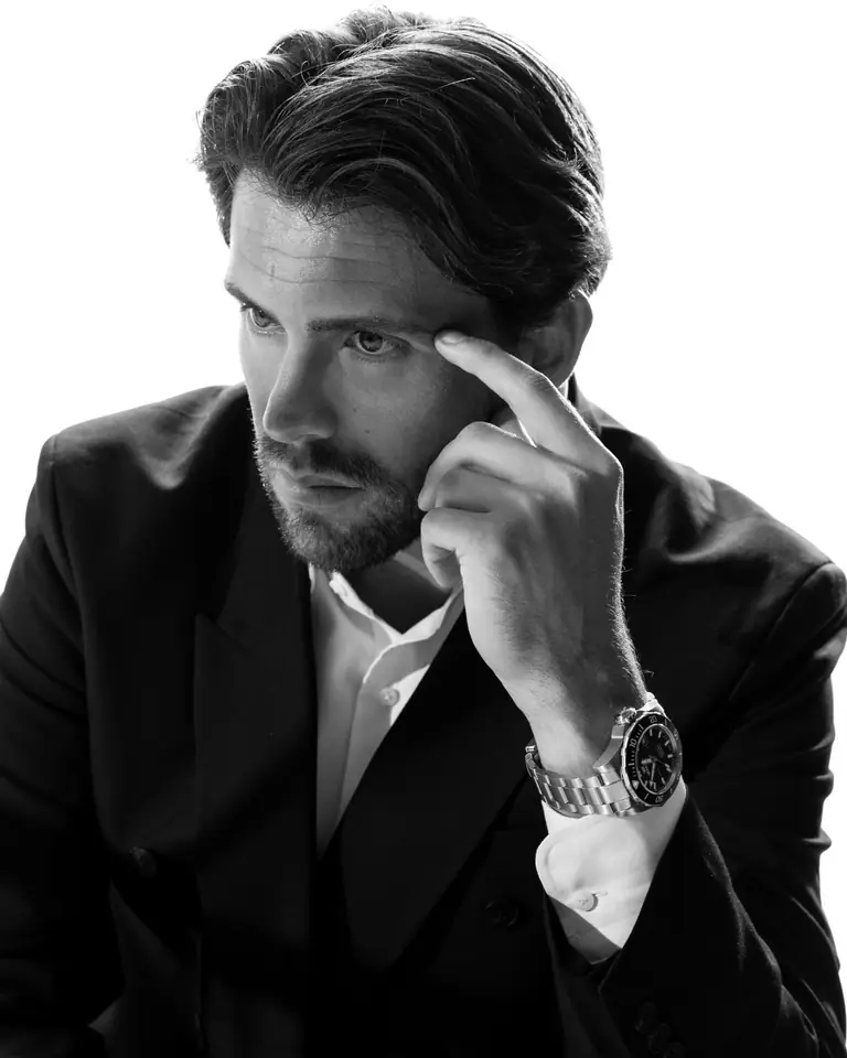 A man in a dark suit wearing a Waldo watch, hand to his temple, in black and white
