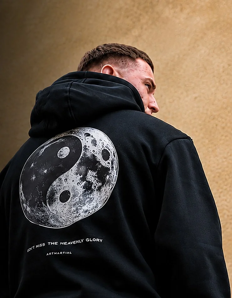 A man in a black ArtMartial hoodie with a moon yin-yang print, seen from behind against a sandstone wall